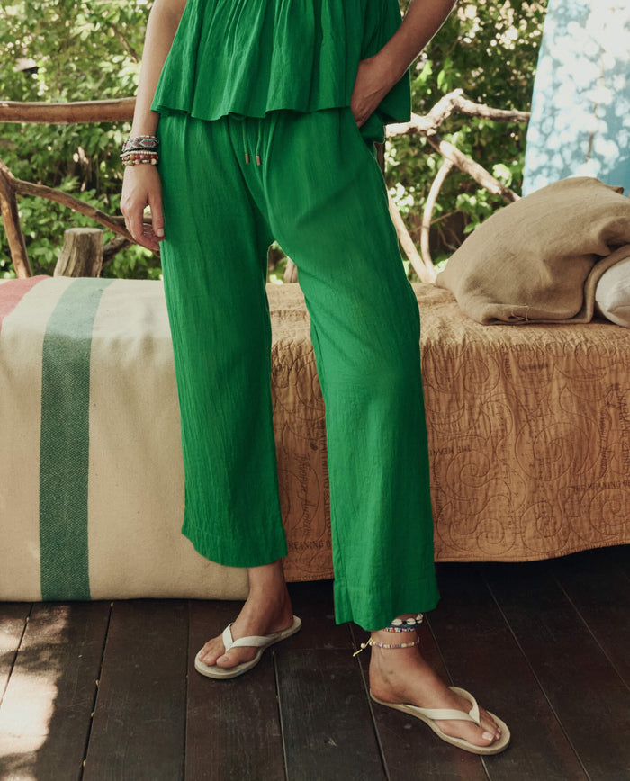 Wearing THE GREAT. SP26 SWIM's The Reef Pant in Bright Sprig, a person stands by a bed with a tan quilt and striped blanket on a wooden deck, surrounded by greenery.