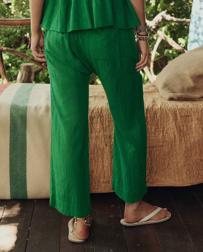 A person stands indoors wearing THE GREAT. SP26 SWIM The Reef Pant in Bright Sprig with a matching green top, white sandals, and layered bracelets, set against a background of a bed and greenery.