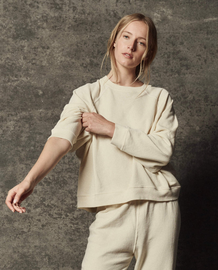 Wearing THE GREAT.'s The Rehearsal Sweatshirt in Washed White, a person stands against a textured gray background, adjusting their sleeve with one hand.