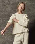 Wearing THE GREAT.'s The Rehearsal Sweatshirt in Washed White, a person stands against a textured gray background, adjusting their sleeve with one hand.