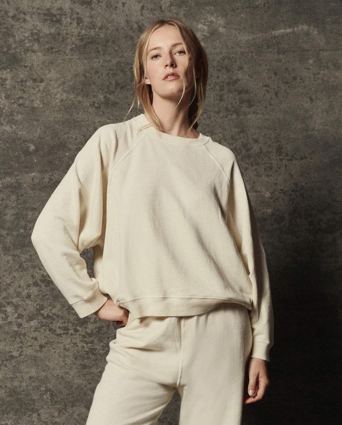 A person wearing THE GREAT.'s The Rehearsal Sweatshirt in Washed White, along with matching pants, stands in front of a textured gray background.