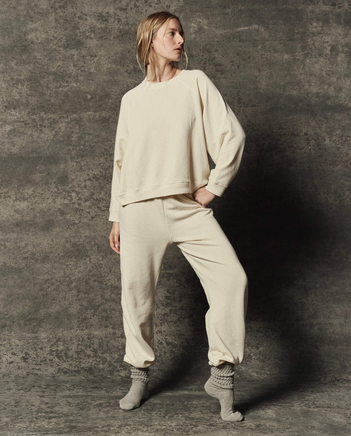 A person stands against a textured background, wearing THE GREAT. PF26 DANCER KNIT BODIES' The Rehearsal Sweatshirt in Washed White, matching sweatpants, and gray socks.