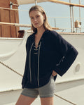 A person wearing THE GREAT. SU26 The Relay Short in Navy Stripe stands in front of a white boat with wooden details and visible ropes.