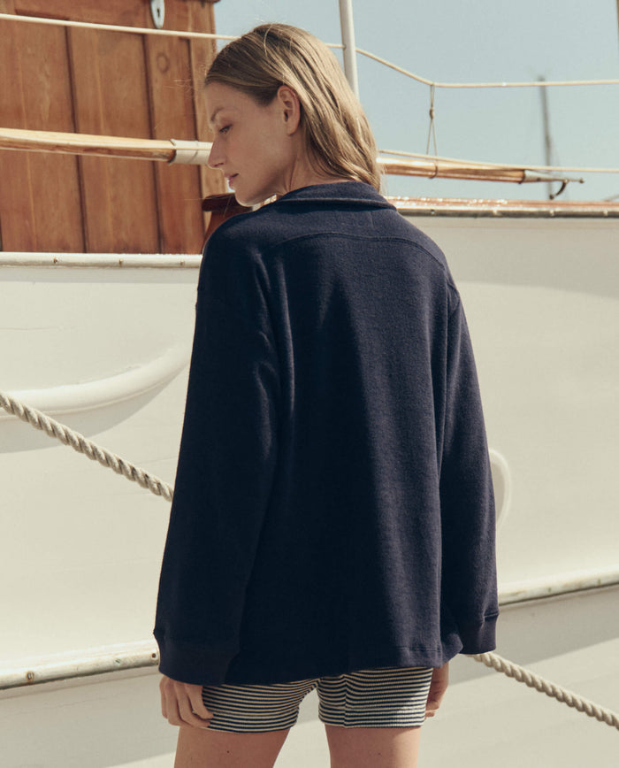 Wearing THE GREAT. SU26's Relay Short in Navy Stripe, a person in a dark long-sleeve top stands on a boat deck facing away, with a wooden structure and rope visible in the background.