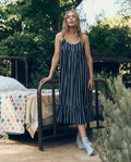 A woman in The Great. SP26 Sleep’s The Ruffle Tank Night Dress in Coastline Stripe stands by an outdoor bed with a quilt, surrounded by trees and greenery.