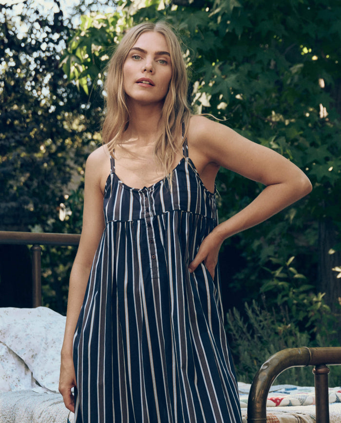 A woman with long blonde hair wears The Ruffle Tank Night Dress in Coastline Stripe by The Great. SP26 Sleep, standing outdoors by a metal bed frame with lush green foliage in the background.