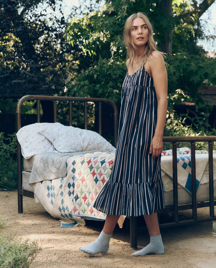 A woman in The Ruffle Tank Night Dress – Coastline Stripe by The Great. SP26 Sleep stands outdoors by a metal-frame bed with quilts and pillows, surrounded by trees and greenery.