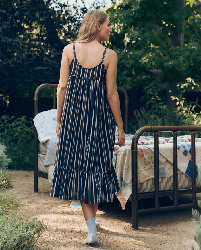 A woman in The Ruffle Tank Night Dress – Coastline Stripe by The Great. SP26 Sleep and socks walks toward an outdoor bed with patchwork quilts in a tranquil garden setting.