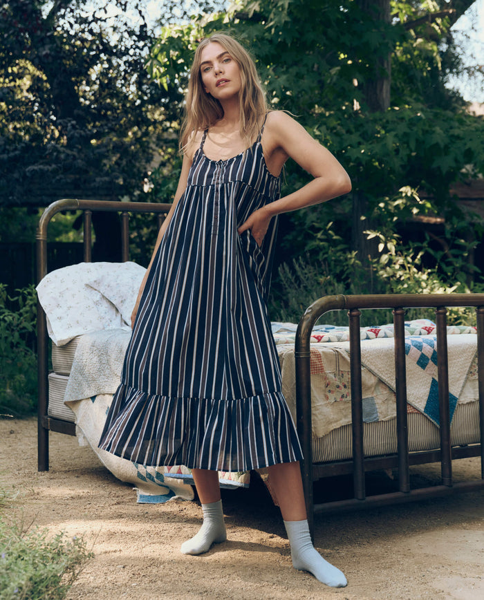 A woman in The Great. SP26 Sleep's The Ruffle Tank Night Dress in Coastline Stripe stands by an outdoor bed with floral and patchwork bedding, surrounded by lush greenery.