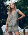A woman wears The Great. SP26 Sleep's Flutter Sleeve Eyelet Top in Bluebell Ditsy, paired with shorts, standing outdoors by a metal bed with quilts amid lush greenery.