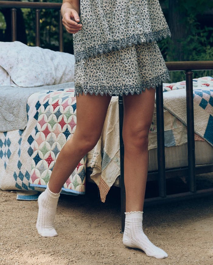A person in a floral dress and white socks stands by a metal bed outdoors, styled with The Great. SP26 Sleep's Smocked Eyelet Short in Bluebell Ditsy for a cozy, stylish touch.