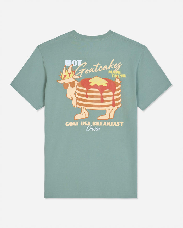 A sage green short-sleeve t-shirt with a retro graphic of a stack of pancakes wearing a crown and sunglasses, with the text 'HOT Goatcakes MADE FRESH' and 'GOAT USA BREAKFAST Union' on a light gray background.
