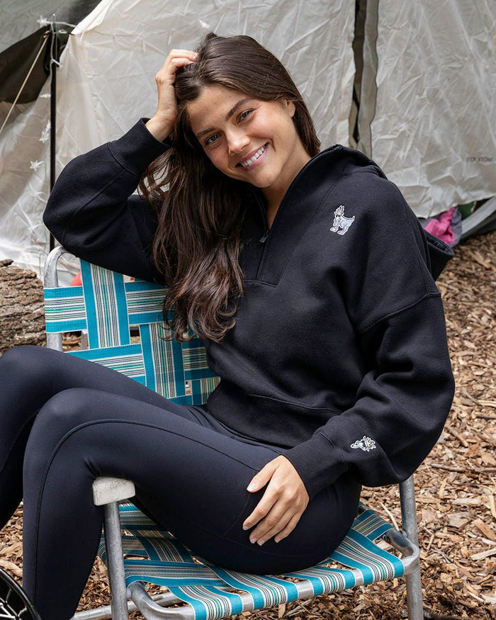 Person wearing a black hoodie with a logo, sitting on chair.#color_black